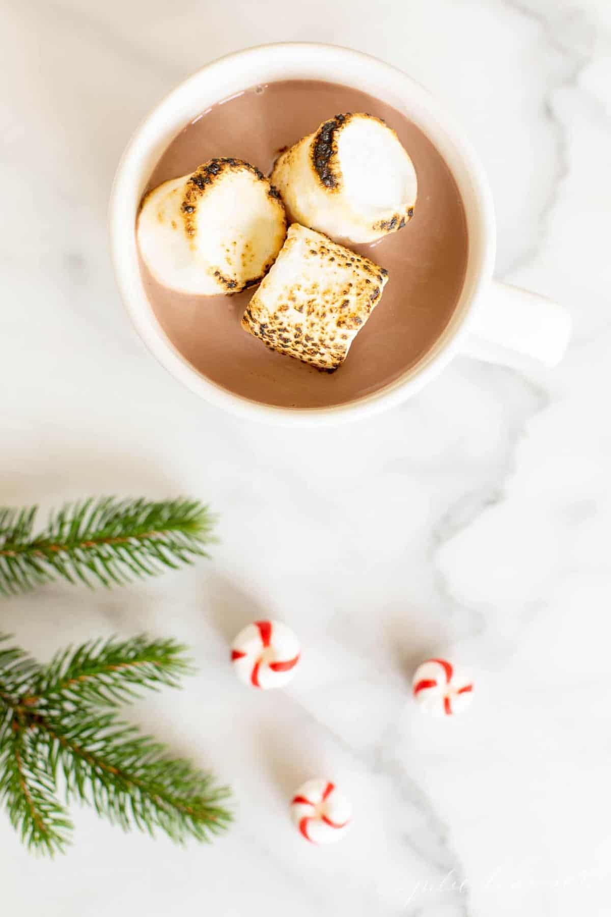 Looking into a mug of peppermint hot chocolate, marshmallows on top, greenery touch and peppermint candies to the side.
