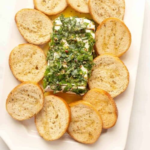 An herb cream cheese recipe surrounded by crostini on a white platter.