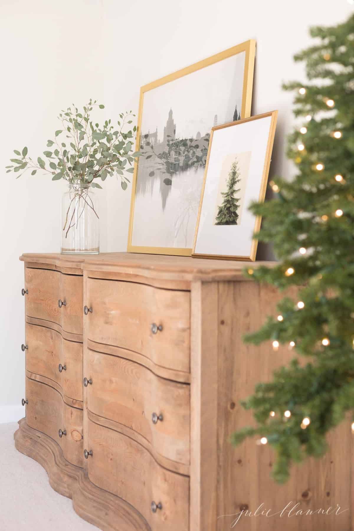 A wooden dresser with a piece of Christmas tree artwork above, tree lights in the foreground of image.