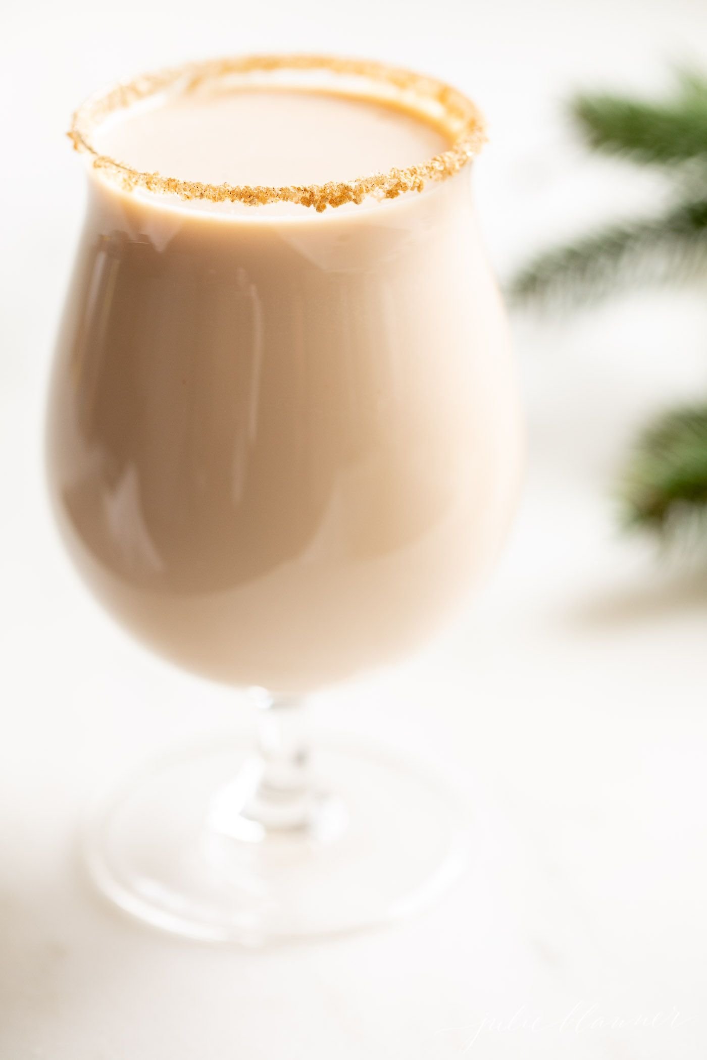 A clear glass with an oatmeal cookie cocktail, fresh winter greens on the edge of the image.