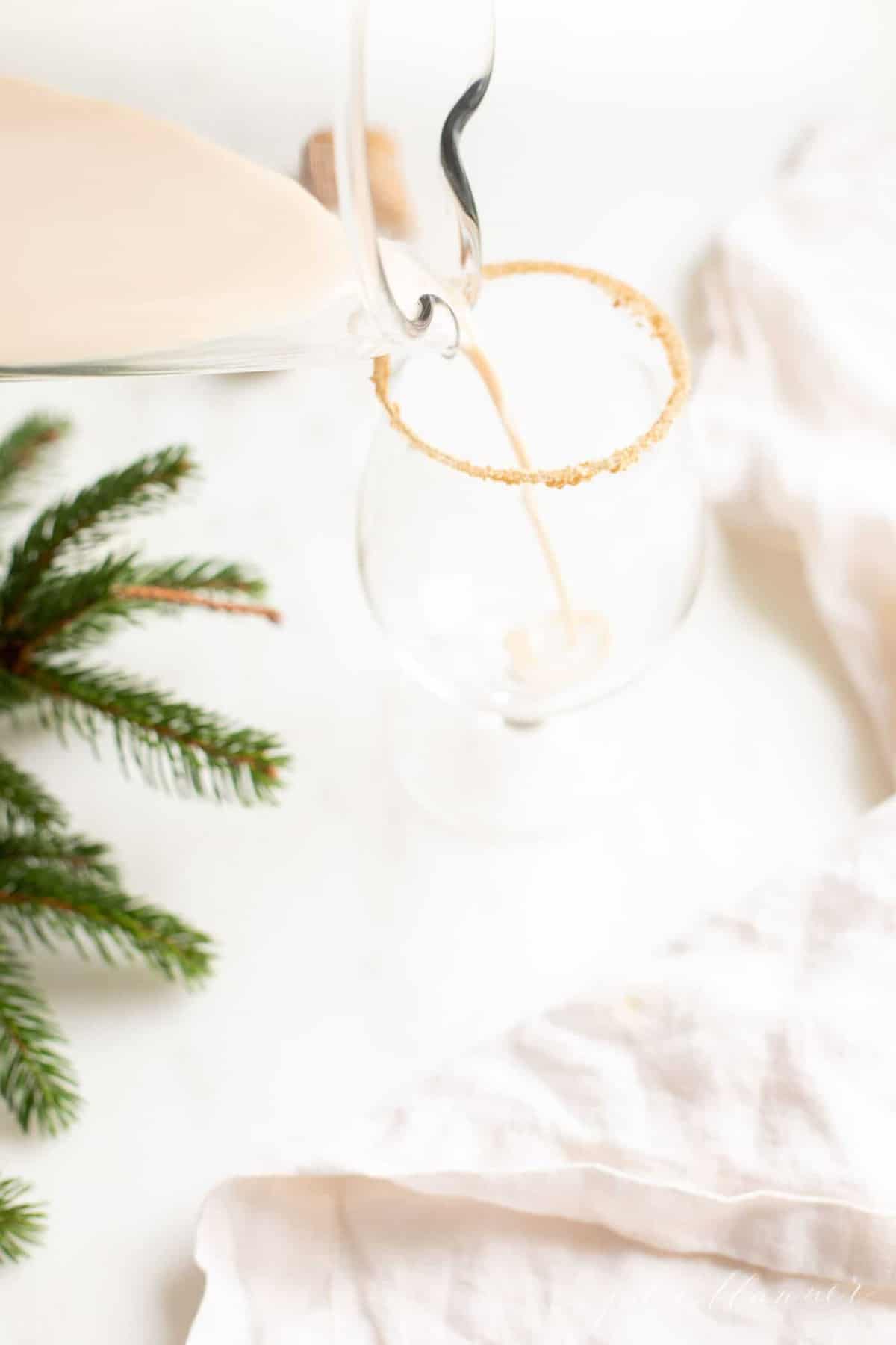 A oatmeal cookie cocktail in a clear glass, holiday greenery to the side, pitcher pouring into glass.