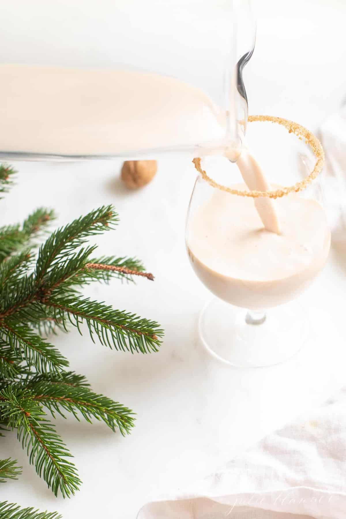 A oatmeal cookie cocktail in a clear glass, holiday greenery to the side, pitcher pouring into glass.