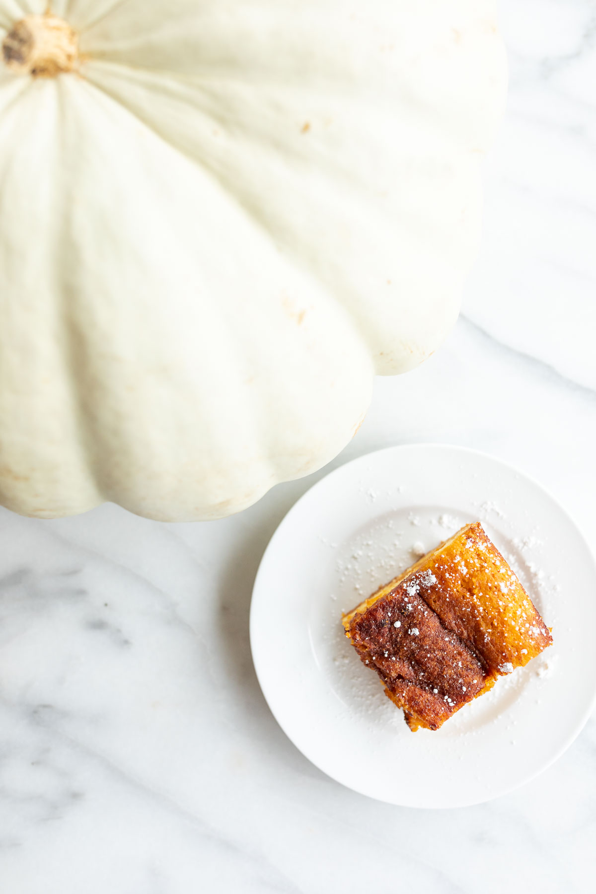 A white plate with a slice of gooey pumpkin butter bars, a white pumpkin beside the plate.