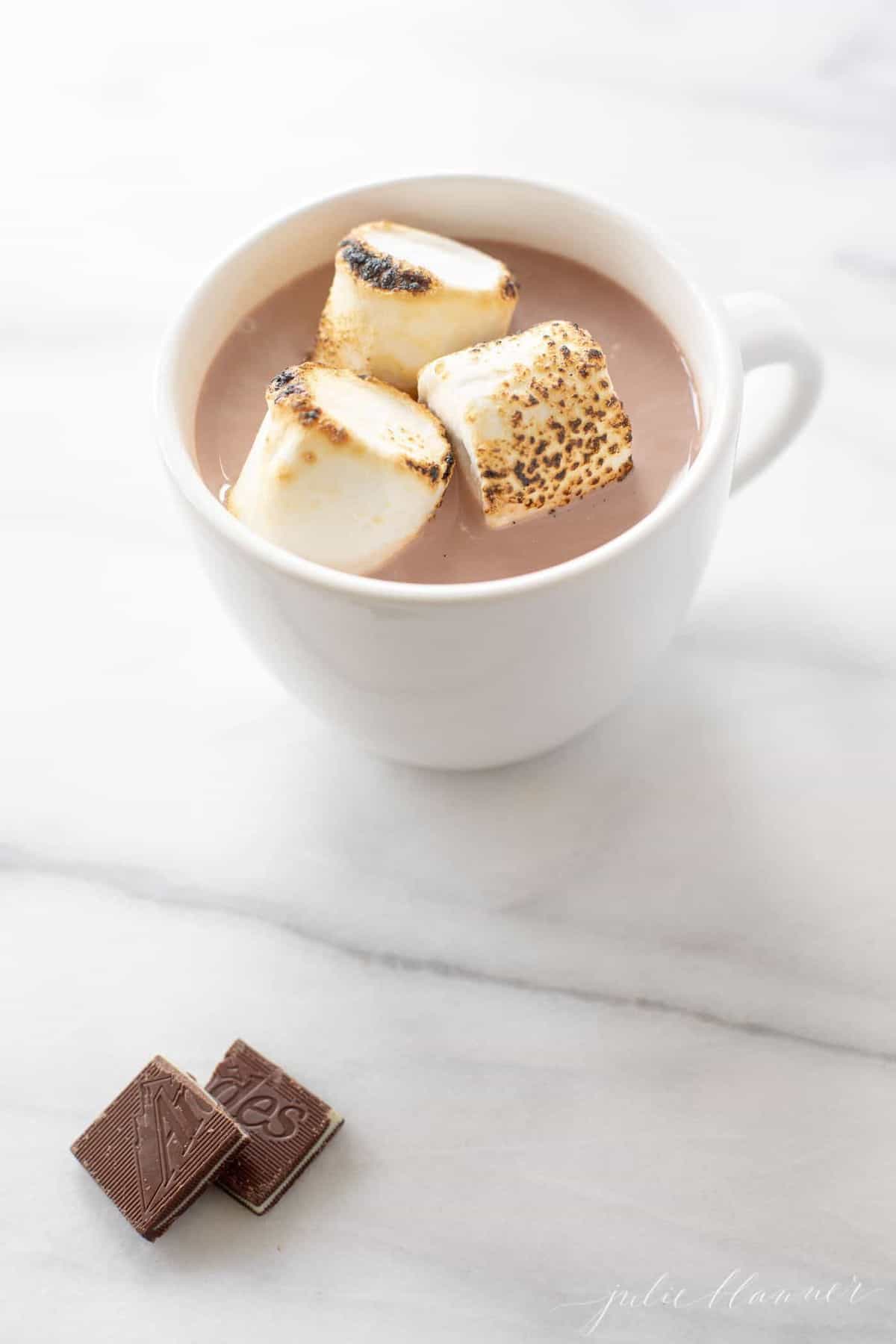 Marble surface, white mug of spiked hot chocolate, marshmallows on top and mint chocolate bar to the side.