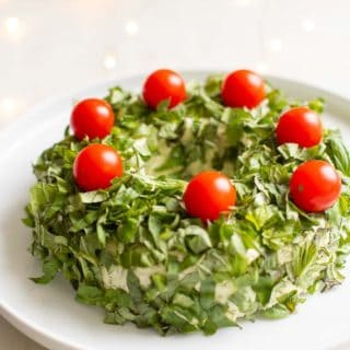 White marble surface with a white platter, featuring a pesto cheese appetizer in the shape of a wreath covered in basil, cheery tomatoes on top for ornaments.