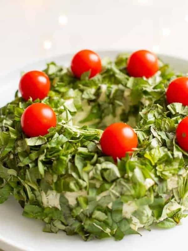 White marble surface with a white platter, featuring a pesto cheese appetizer in the shape of a wreath covered in basil, cheery tomatoes on top for ornaments.