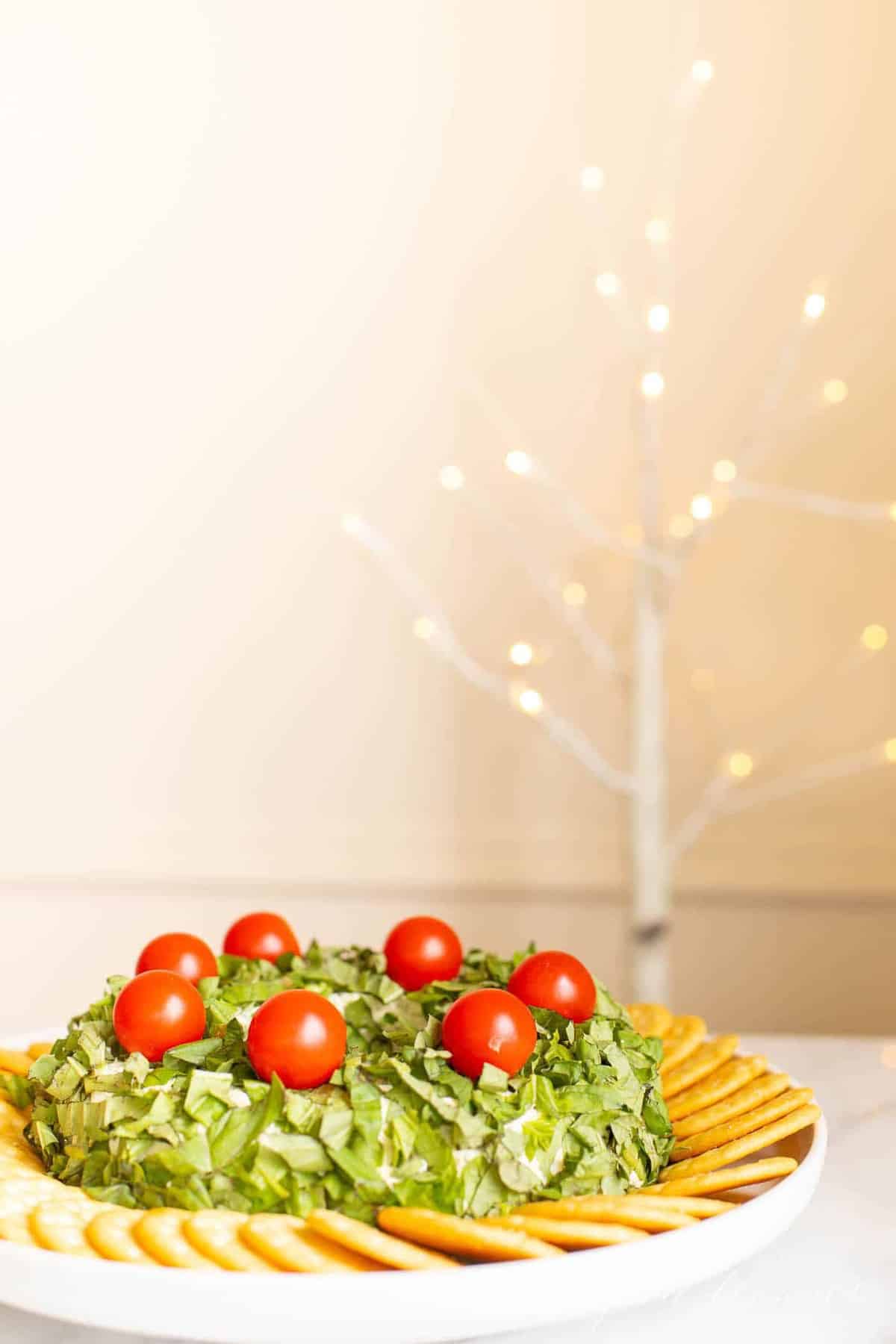 A pesto cheeseball Christmas wreath appetizer surrounded by crackers on a platter. Sparkling tree in the background.