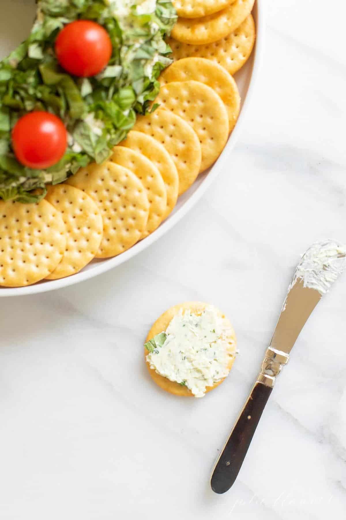 Edge of a platter with a pesto cheese appetizer, crackers surrounding. Single cracker with cheese on top and knife to the side.