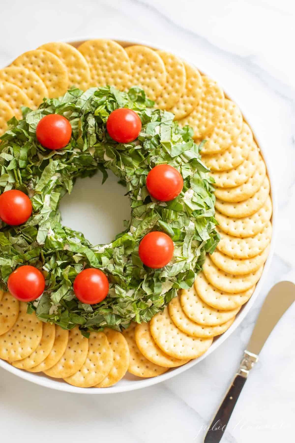 A pesto cheeseball Christmas wreath appetizer surrounded by crackers on a platter, knife to the side.