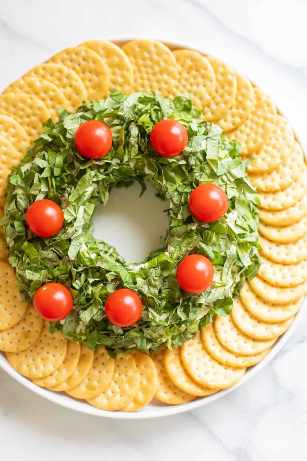 A pesto cheeseball Christmas wreath appetizer surrounded by crackers on a platter.