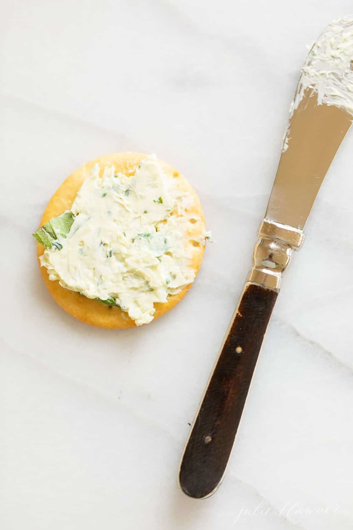 Marble surface with a single cracker covered in pesto cheeseball spread, knife to the side.
