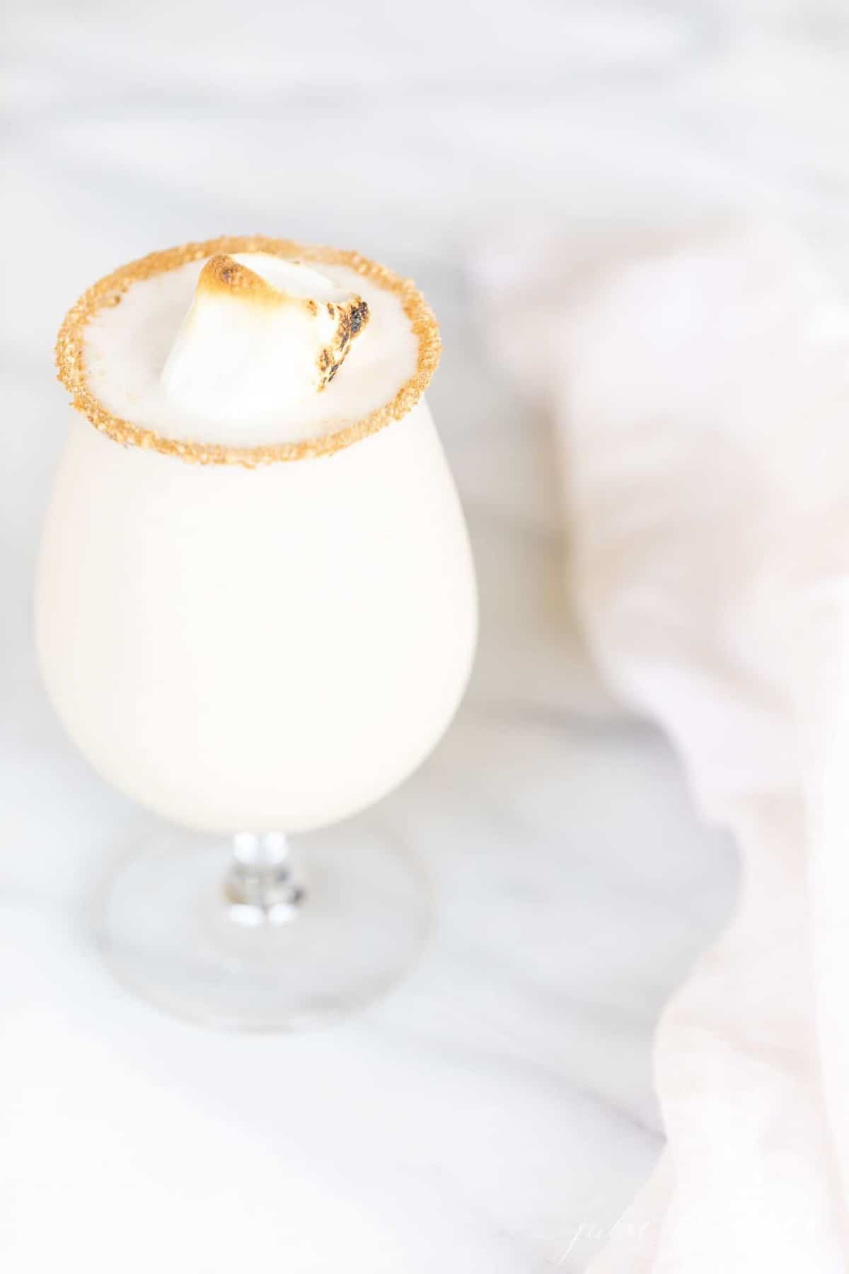 A marble counter top with a clear glass featuring an amaretto liqueur snowball recipe, toasted marshmallow for garnish.