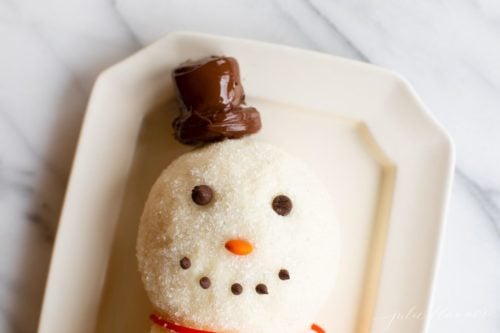 White surface, snowman cheeseball on a platter, chocolate chip smile, chocolate marshmallow hat.