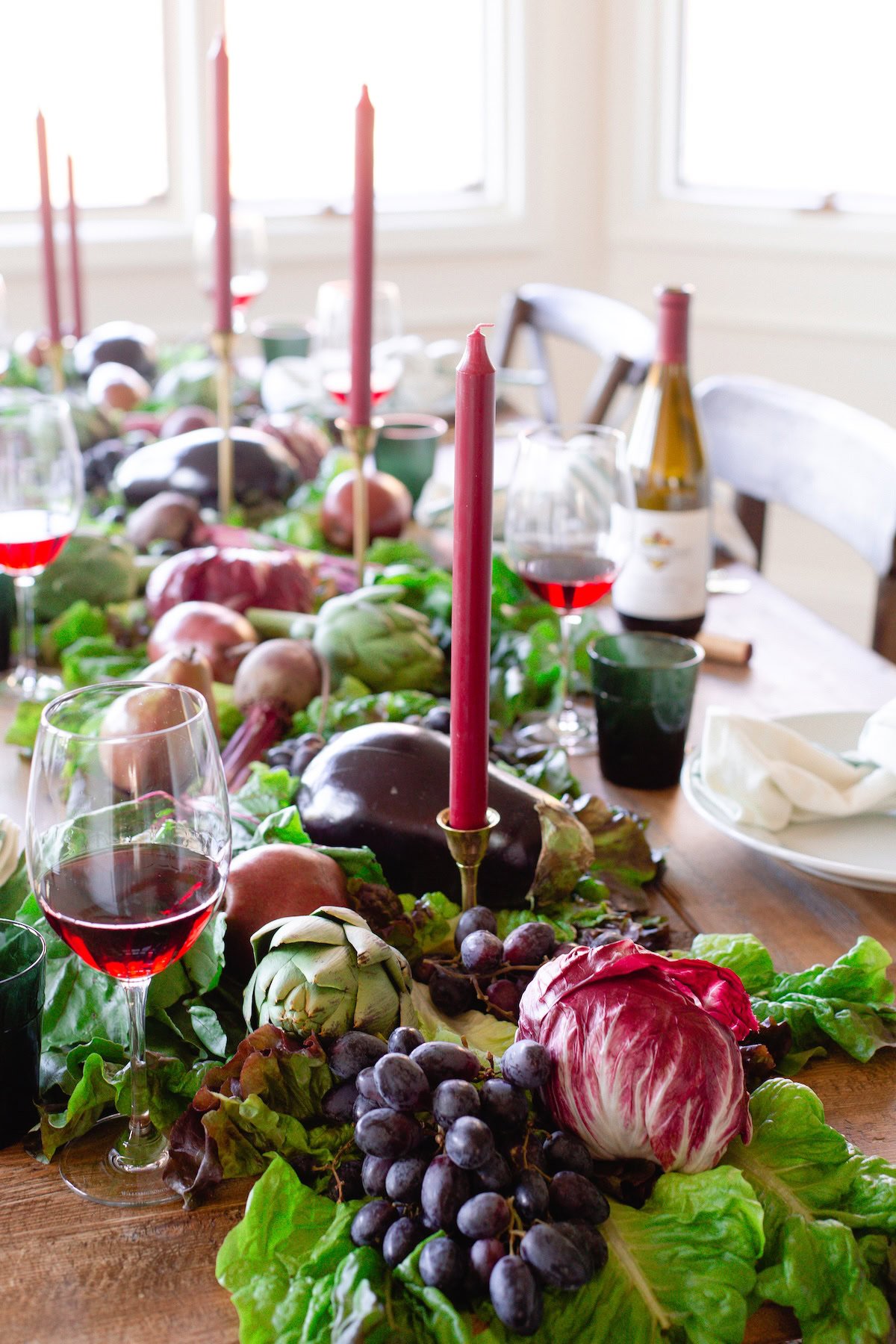 A wooden dining table set with wine glasses, a wine bottle, red candles, and a vibrant centerpiece of fresh vegetables and grapes on leafy greens is the perfect inspiration for How to Host Thanksgiving in style.