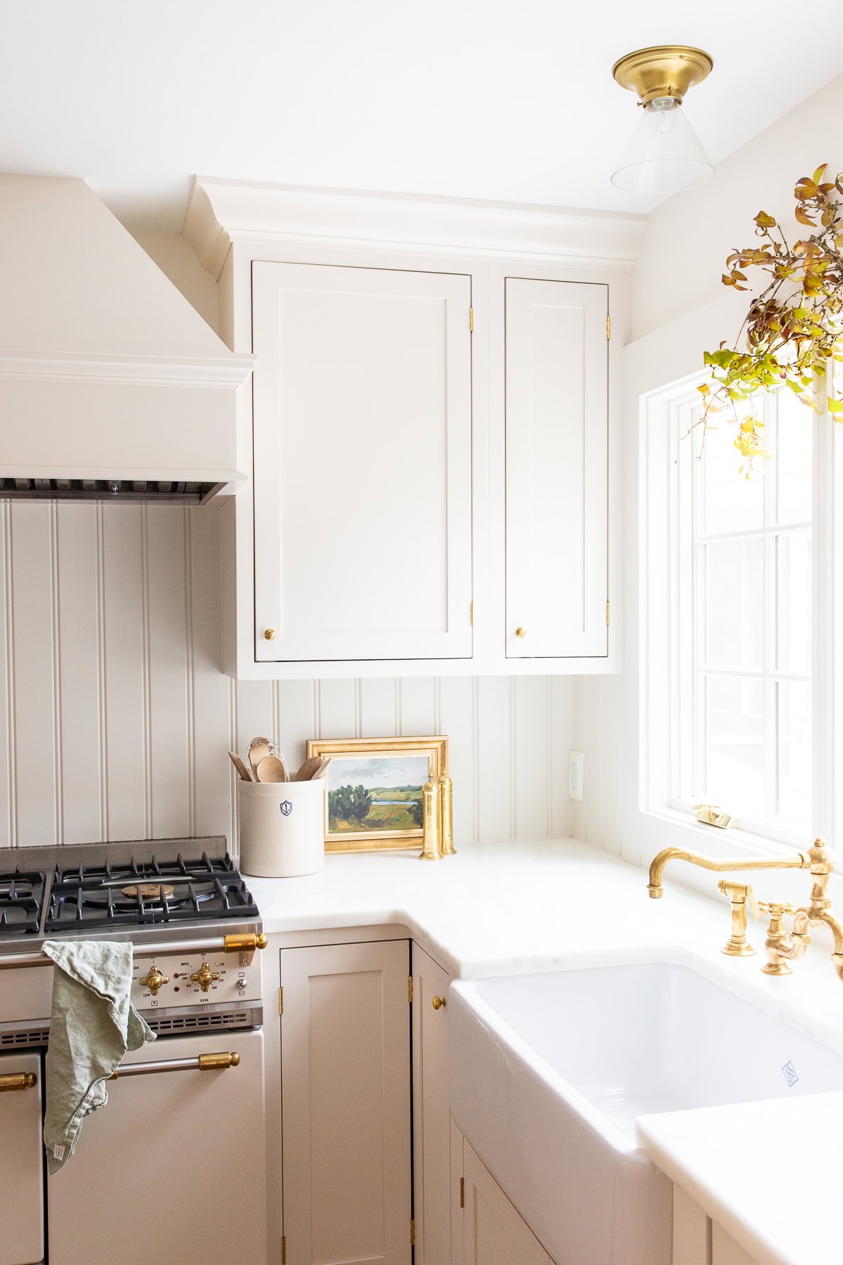Bright kitchen with white cabinets, gold hardware, a farmhouse sink, and a gas stove—perfect for learning How to Host Thanksgiving. Natural light streams through the window; a small painting and utensils decorate the counter.