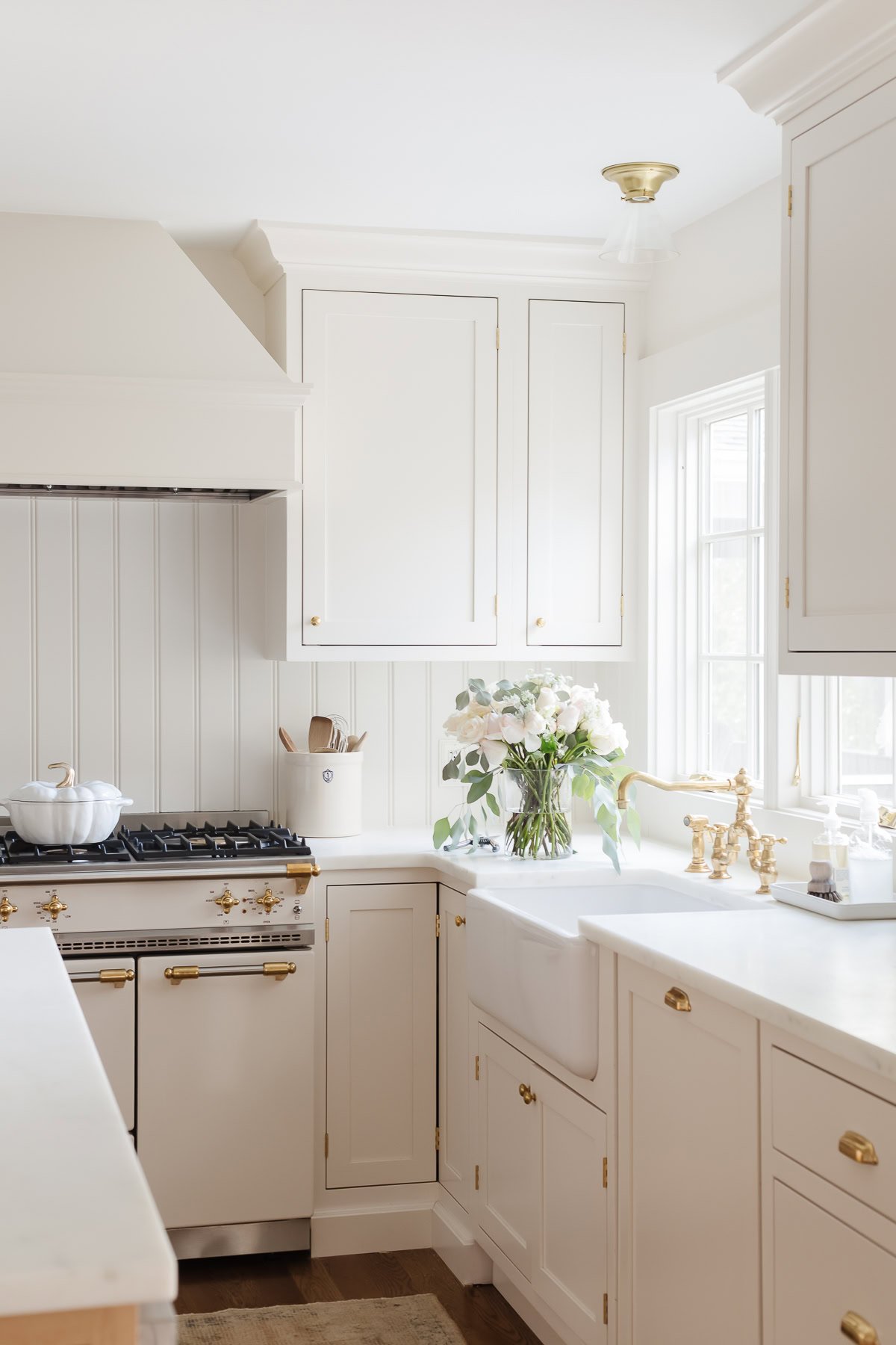 Bright kitchen with white cabinets, gold hardware, farmhouse sink, and gas stove—perfect for learning how to host Thanksgiving. A vase of white flowers sits on the counter near a window, adding a festive touch.