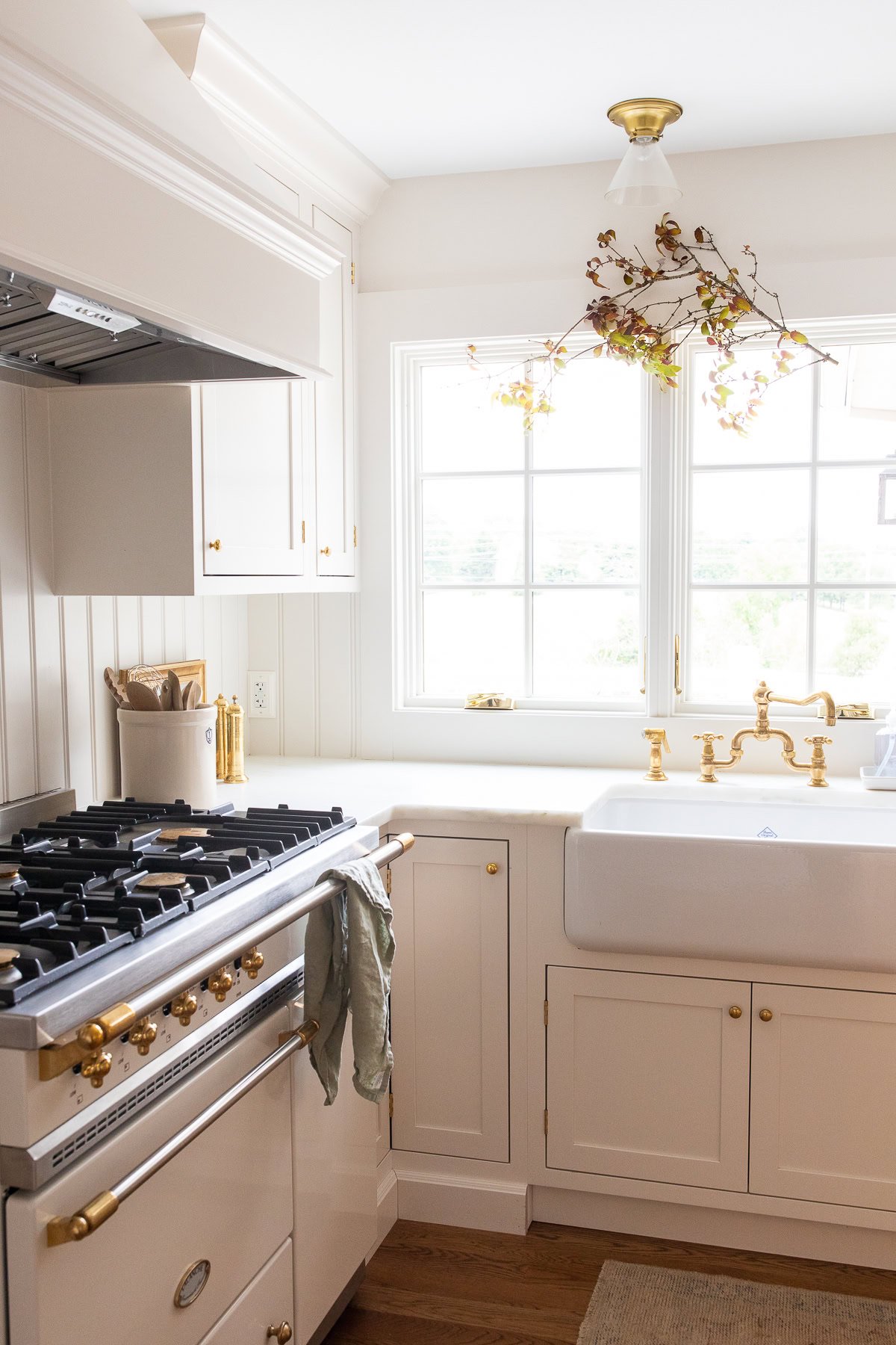 A bright kitchen with a gas stove, white cabinets, farmhouse sink, gold fixtures, and a branch with leaves above the window—an ideal setting for learning how to host Thanksgiving in style.
