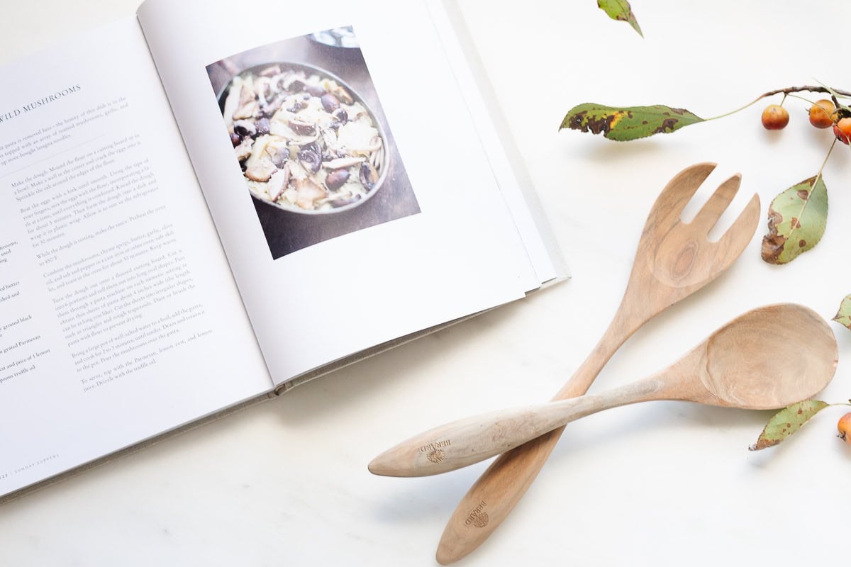 Open cookbook with a mushroom dish photo, wooden salad fork and spoon beside it, and some leafy branches with small fruits on a white surface—perfect inspiration for How to Host Thanksgiving.