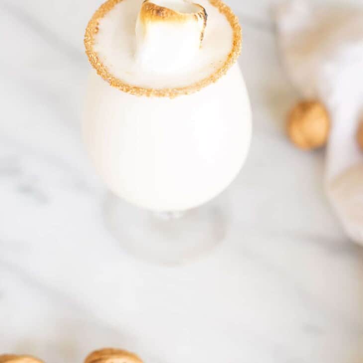 A marble counter top with a clear glass featuring a snowball recipe, toasted marshmallow for garnish.