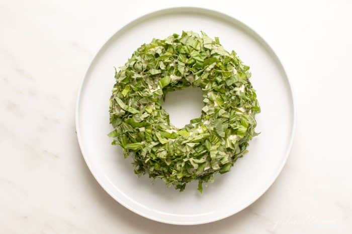 White marble surface with a white platter, featuring a pesto cheese appetizer in the shape of a wreath covered in basil.