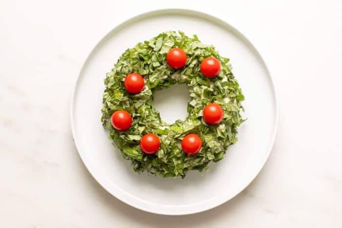 White marble surface with a white platter, featuring a pesto cheese appetizer in the shape of a wreath covered in basil.