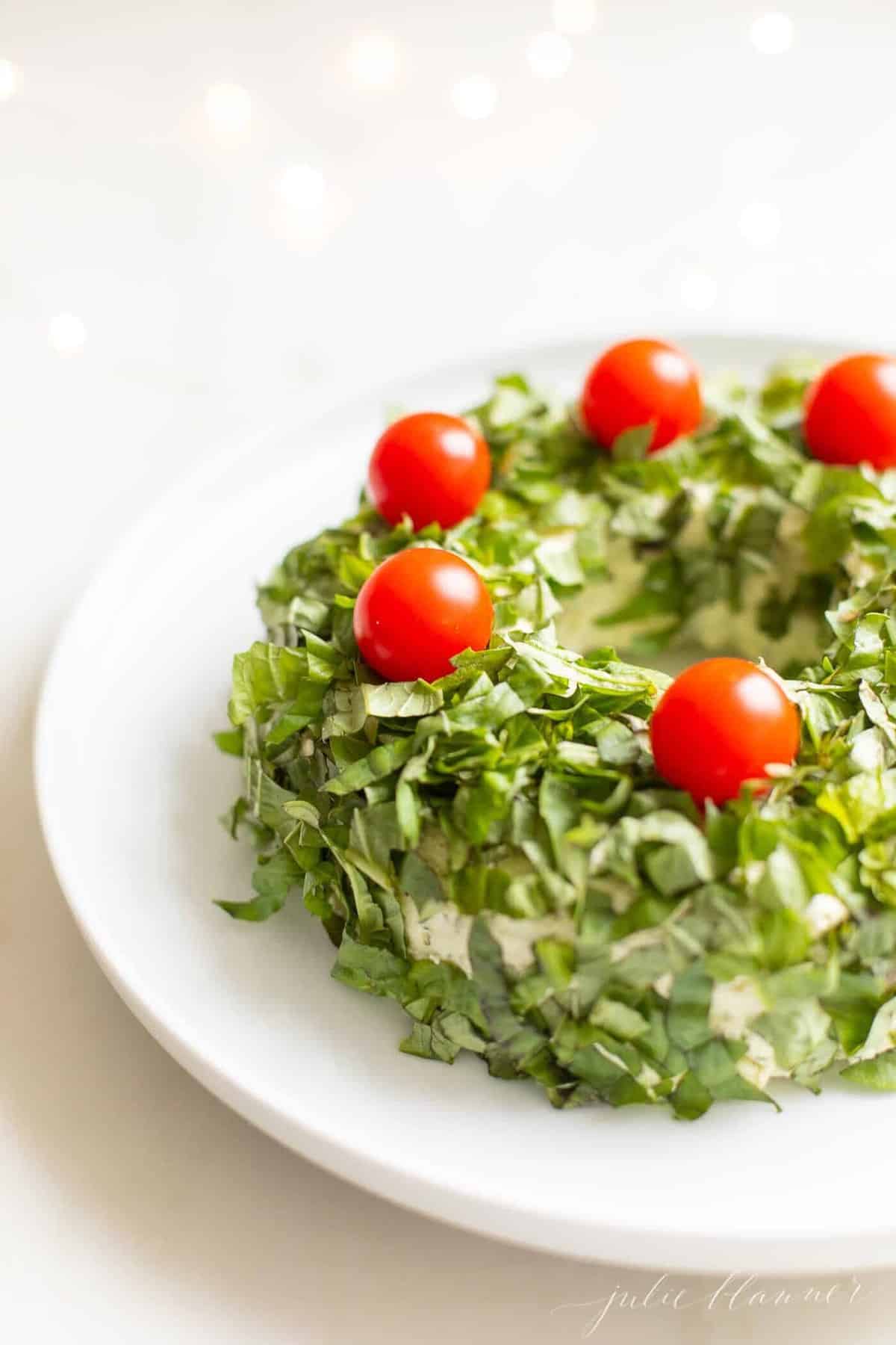 White marble surface with a white platter, featuring a pesto cheese appetizer in the shape of a wreath covered in basil, cheery tomatoes on top for ornaments.