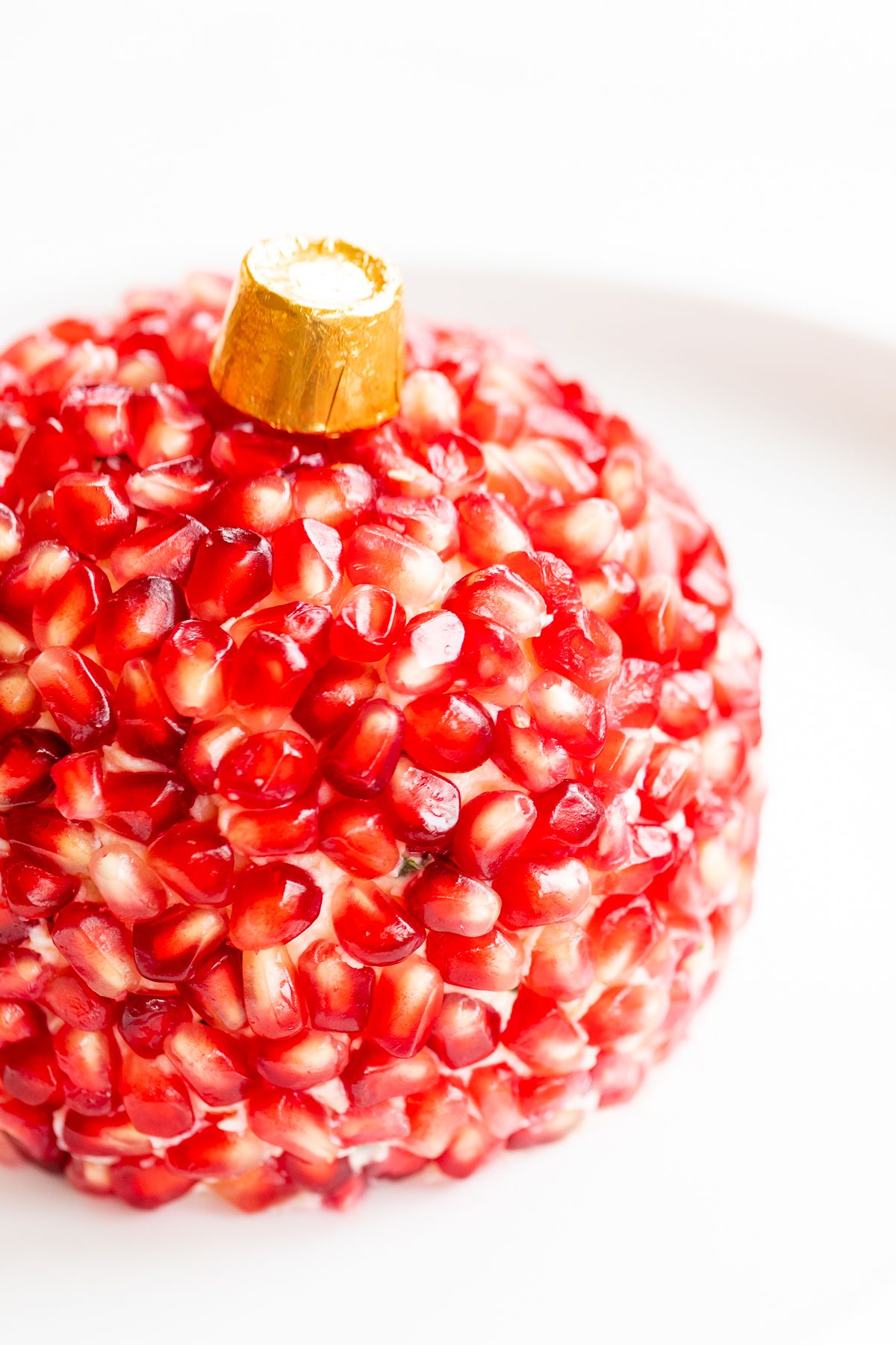 A pomegranate Christmas cheese ball shaped into an ornament on a round white plate.