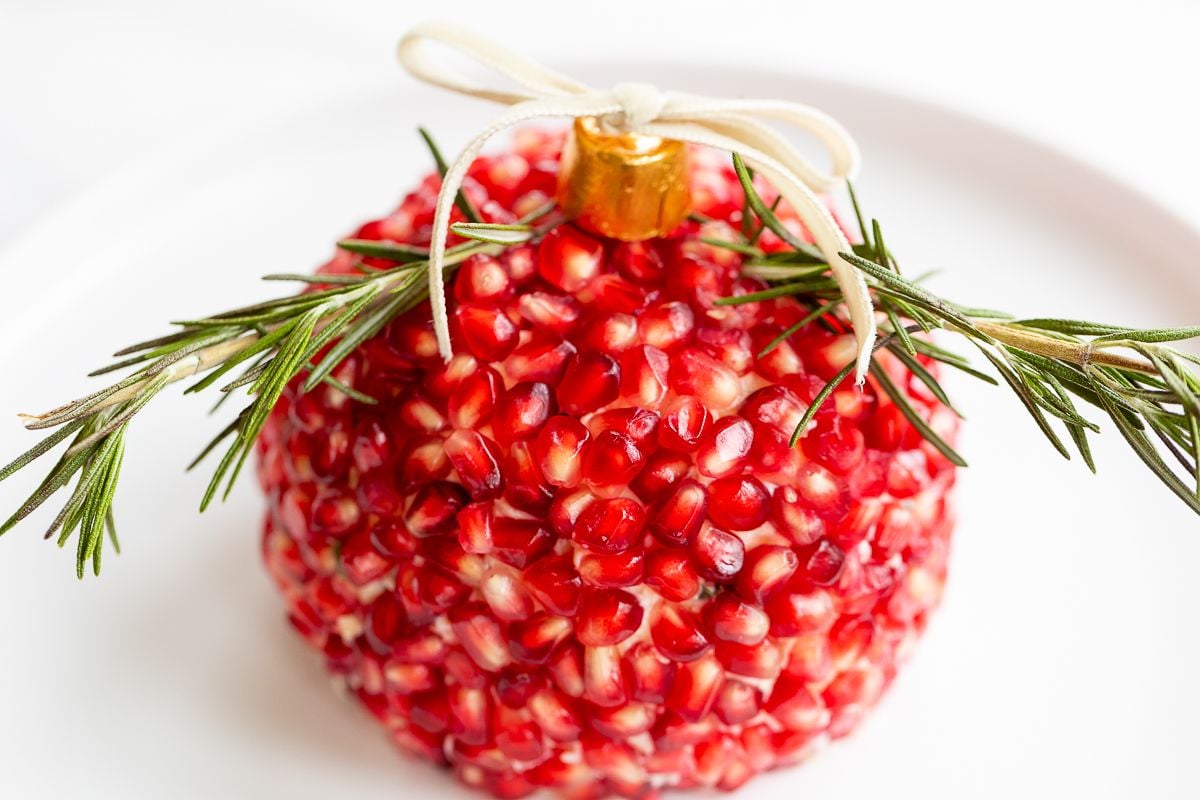 A pomegranate Christmas cheese ball shaped into an ornament on a round white plate.