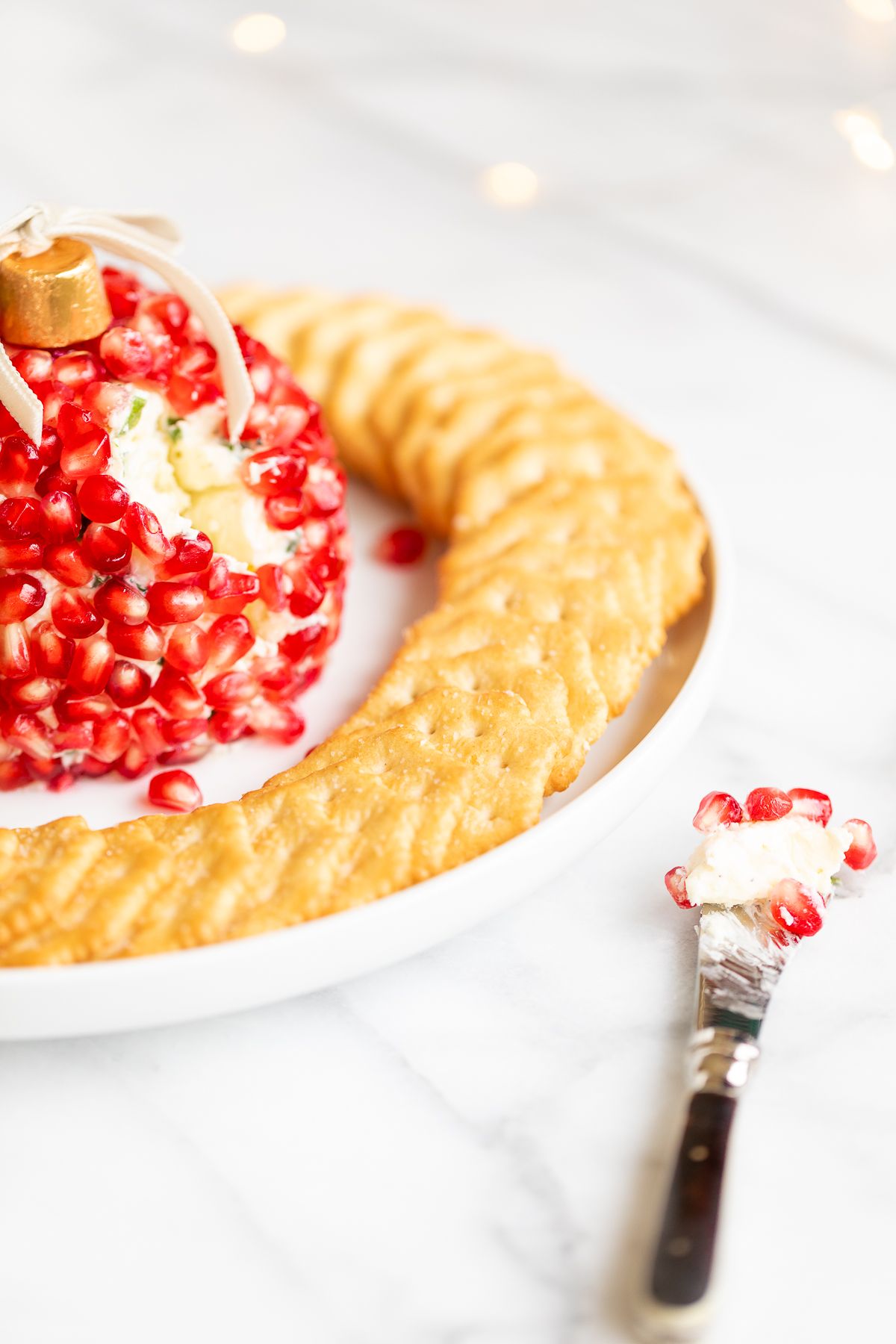 A pomegranate Christmas cheese ball shaped into an ornament, surrounded by crackers on a round white plate.