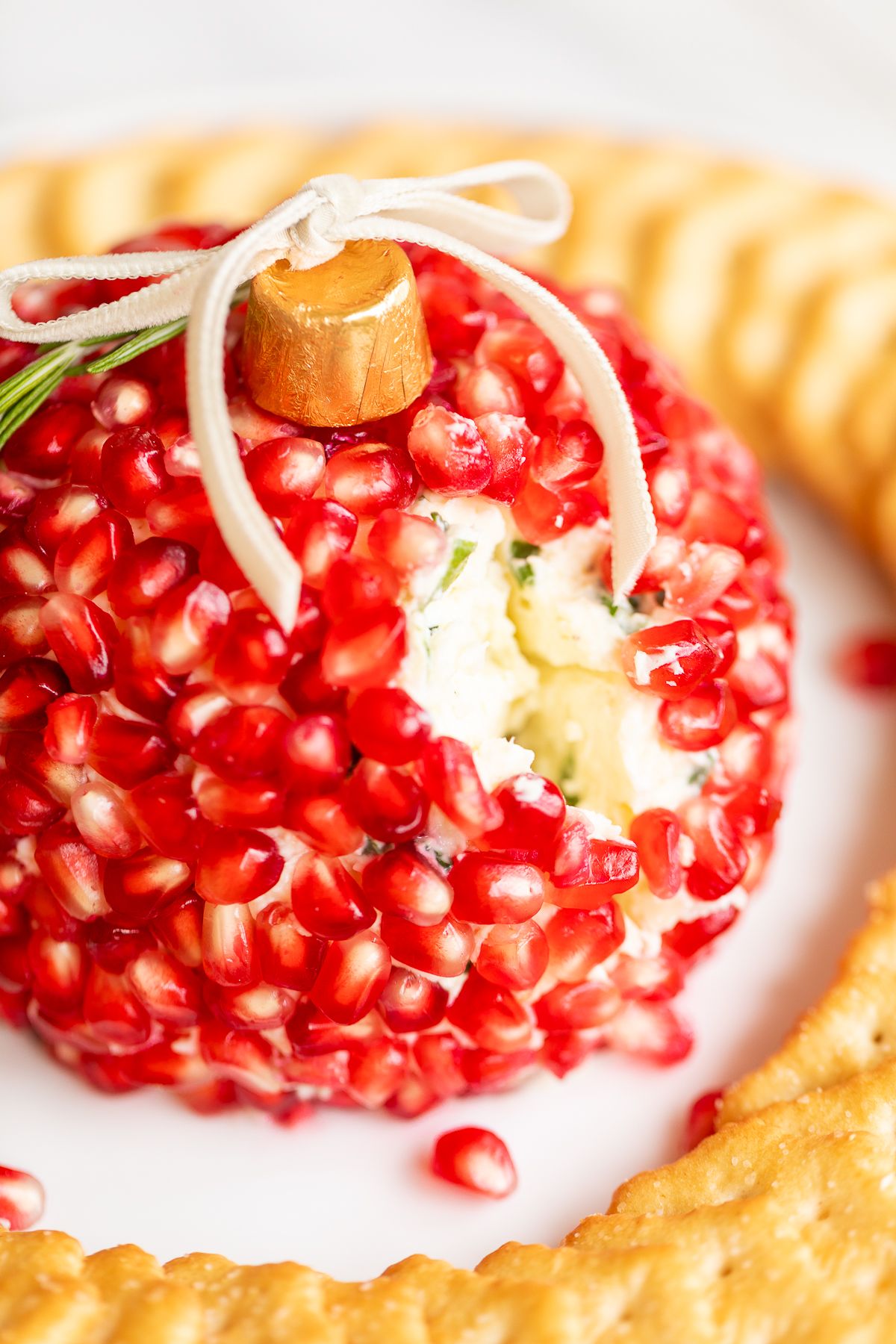 A pomegranate Christmas cheese ball shaped into an ornament, surrounded by crackers on a round white plate.