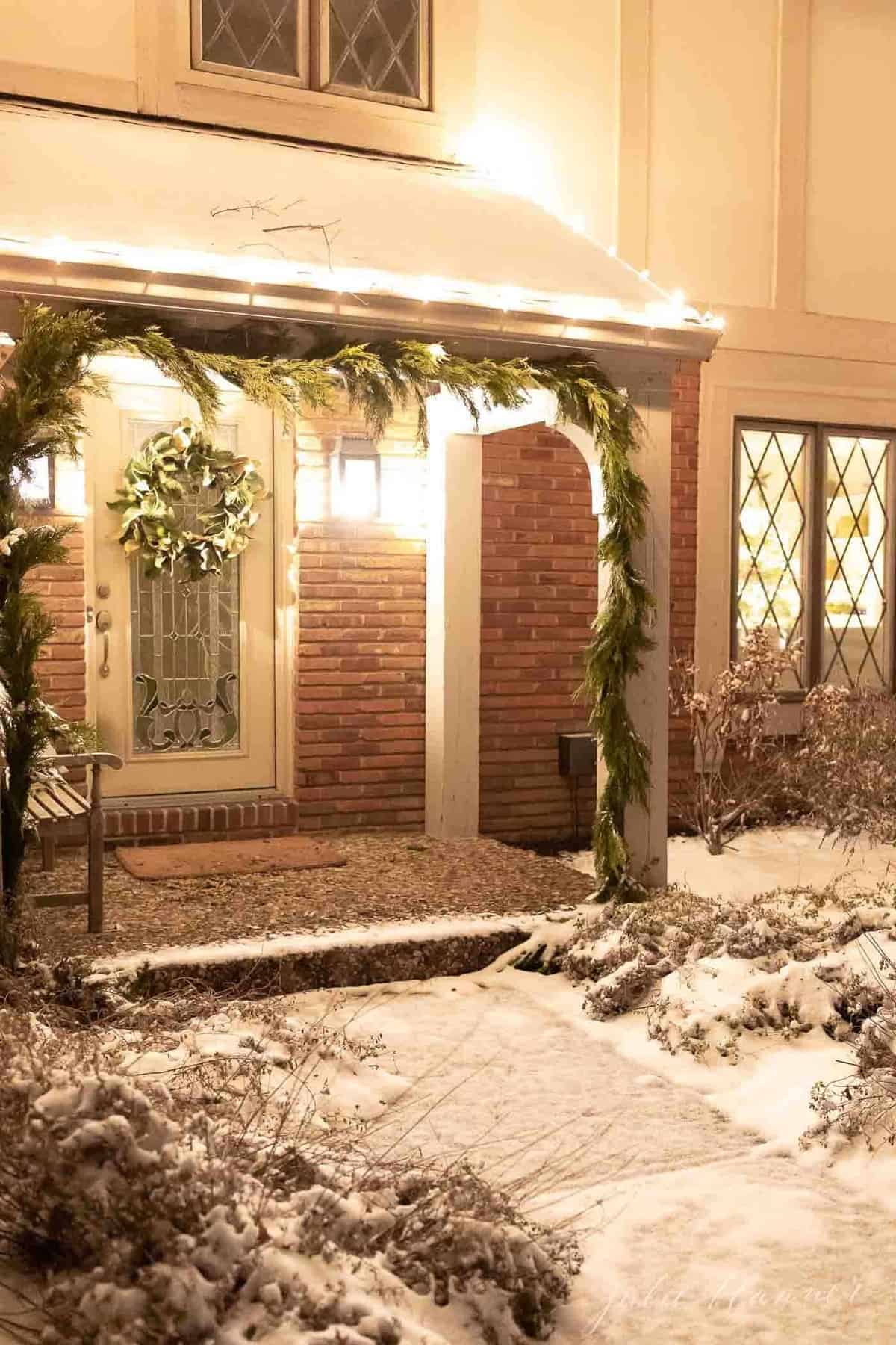 Exterior of a home, snow on the ground and greenery garland on posts for Christmas.