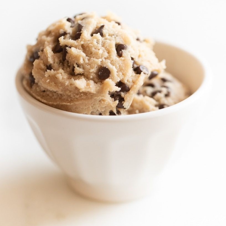 A small white bowl full of edible cookie dough with chocolate chips, on a marble surface