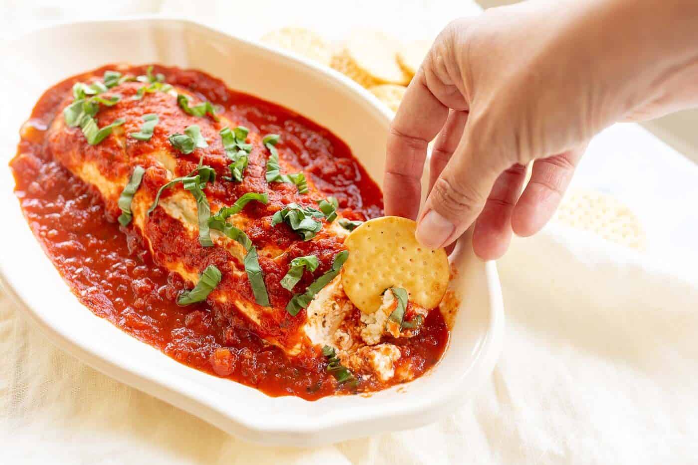 A single white serving dish filled with baked goat cheese covered in marinara, hand reaching in with a cracker..