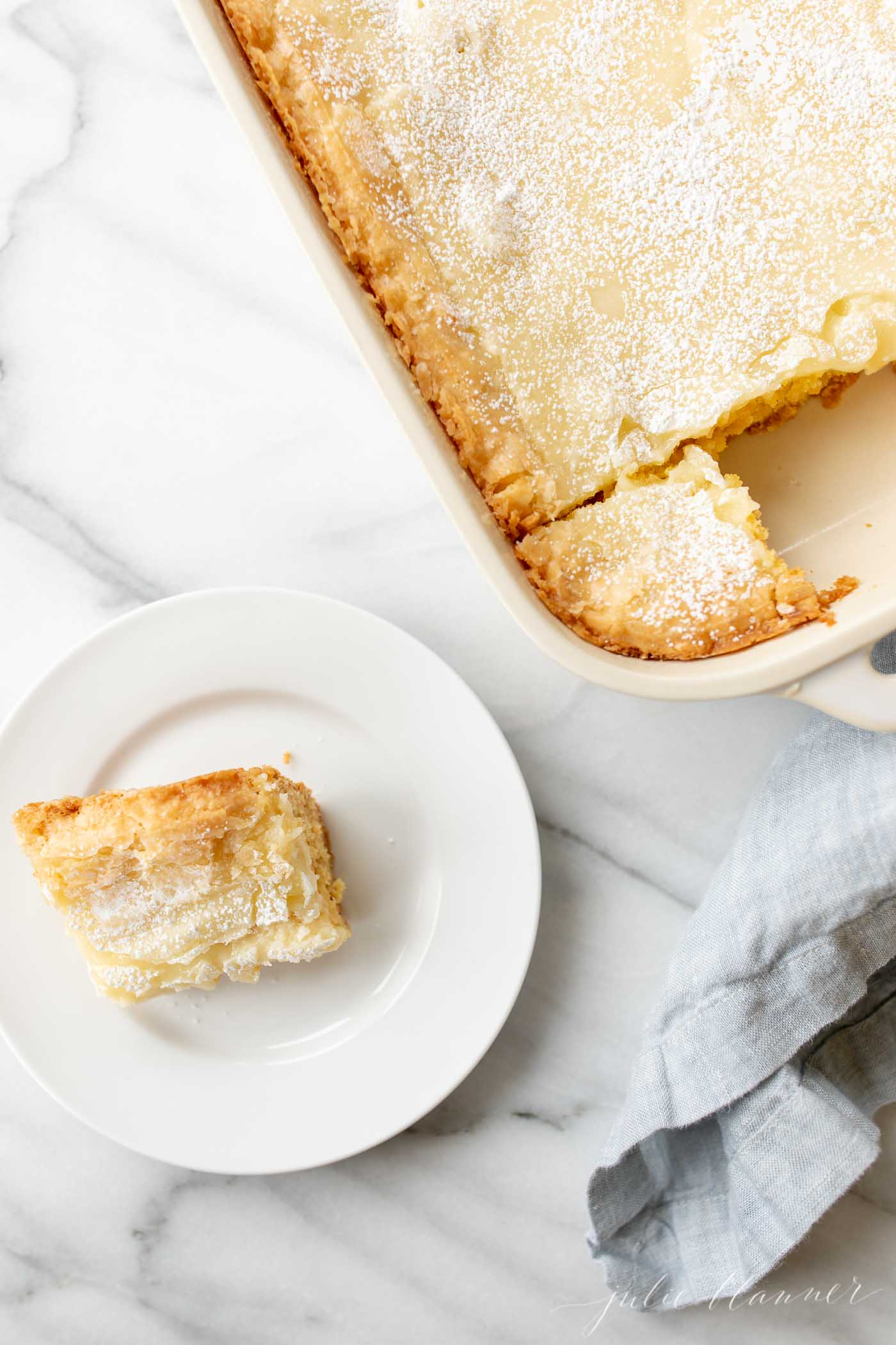 A white rectangular baking dish filled with a homemade gooey butter cake. White plate with a serving to the side.