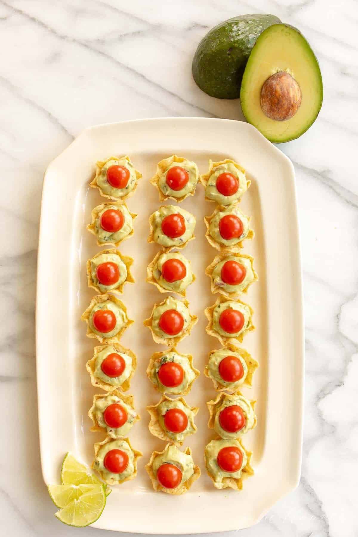 A platter filled with chips, served as guacamole bites, topped with a slice of cherry tomato, avocado to the side.