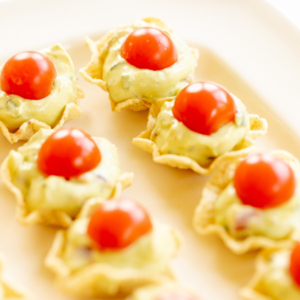 A white tray full of guacamole bites made from tortilla chips, topped with sliced cherry tomatoes.