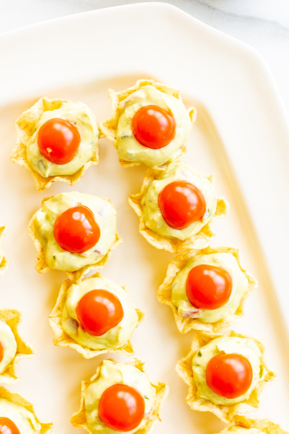 A white tray full of guacamole bites made from tortilla chips, topped with sliced cherry tomatoes.