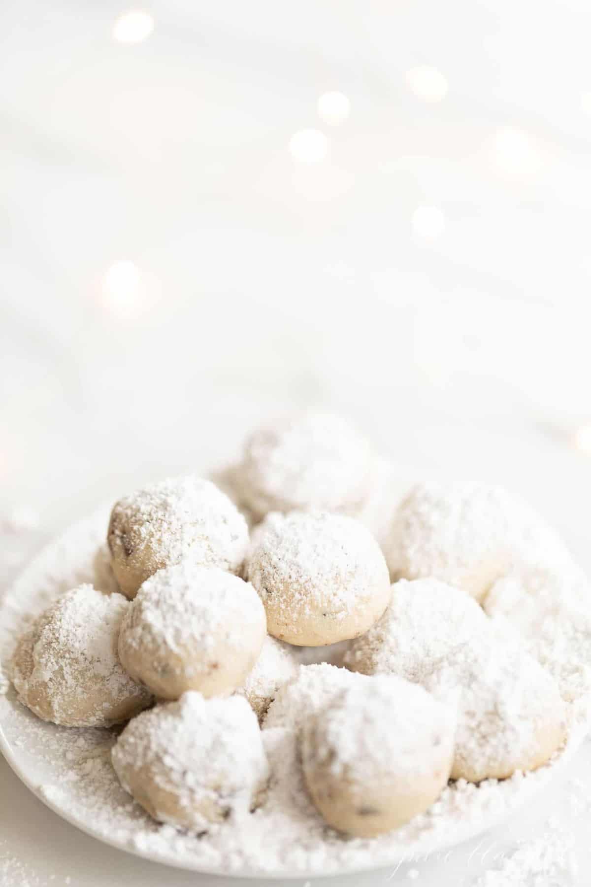 A white plate on a white surface featuring snowball cookies dusted in powdered sugar.