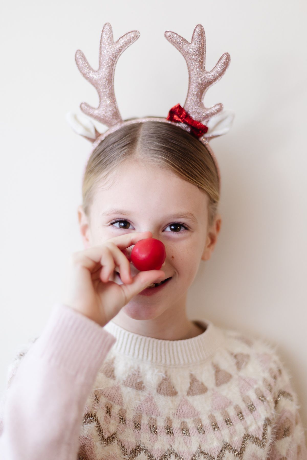 A little girl in reindeer antlers holding a red no bake Rudolph nose Christmas treat up to her nose.