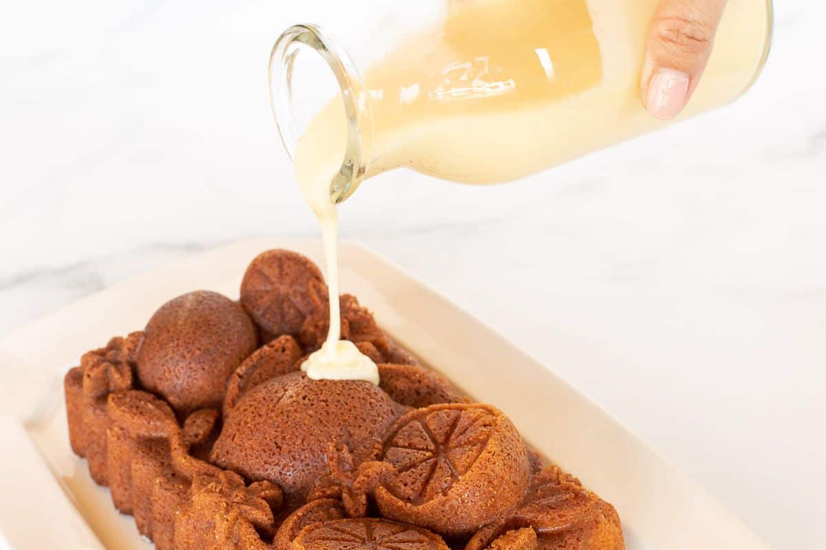 An orange pound cake shaped from a citrus loaf pan, on a white platter, as a glass carafe of glaze is being poured on top.