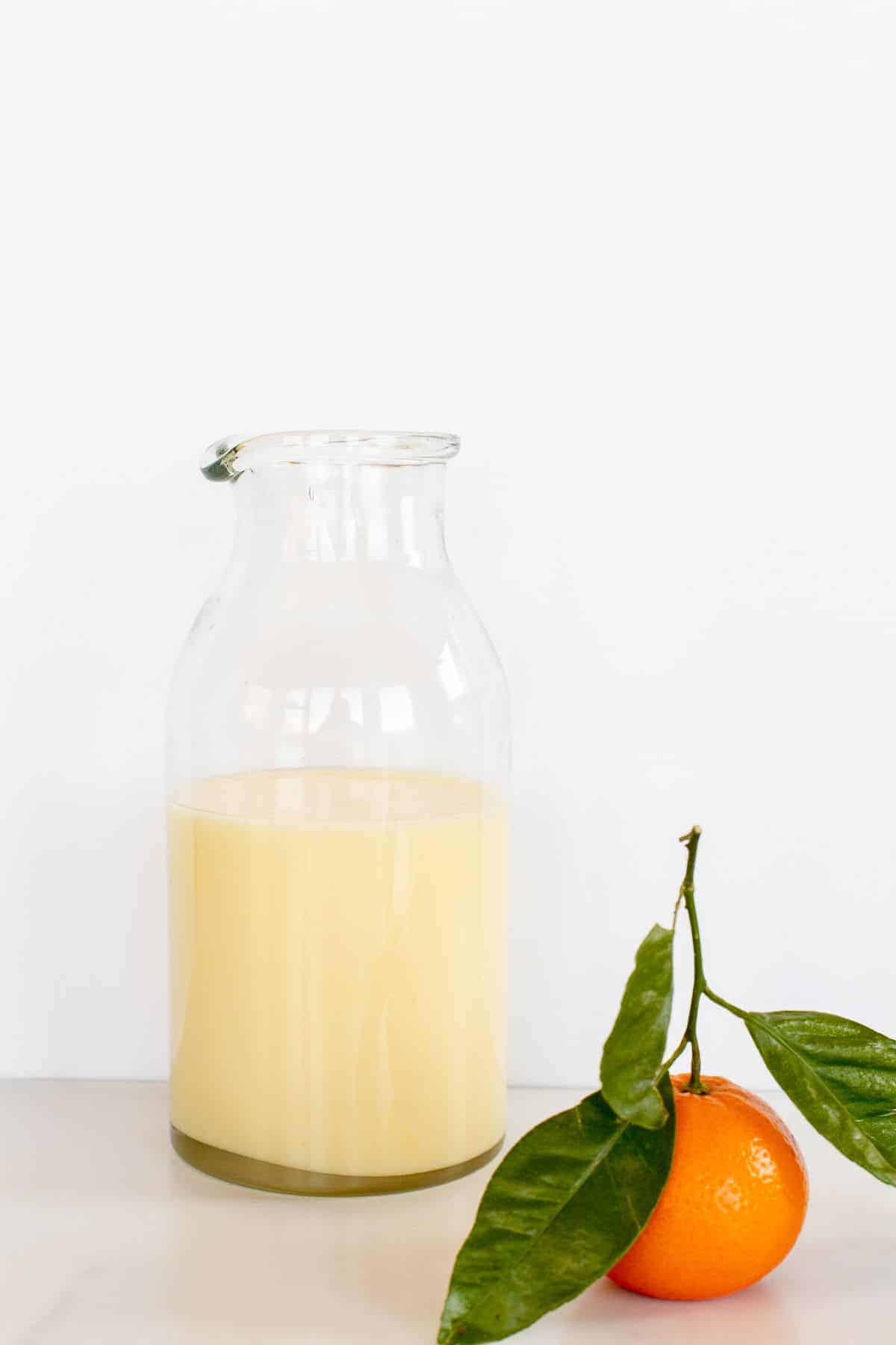 A glass container full of orange glaze, with a fresh orange resting next to it on a marble surface.