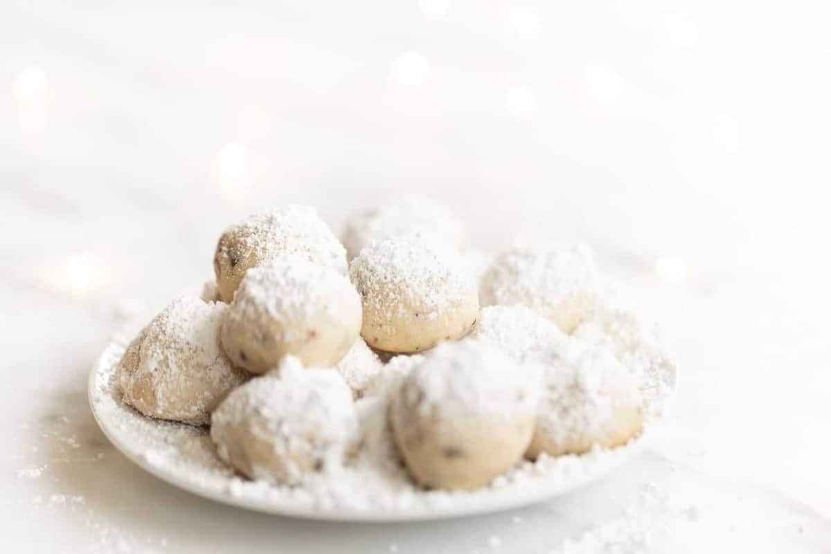 A white plate on a white surface featuring snowball cookies dusted in powdered sugar.