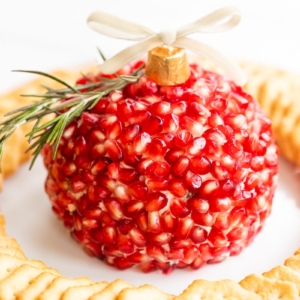 A pomegranate Christmas cheese ball shaped into an ornament, surrounded by crackers on a round white plate.