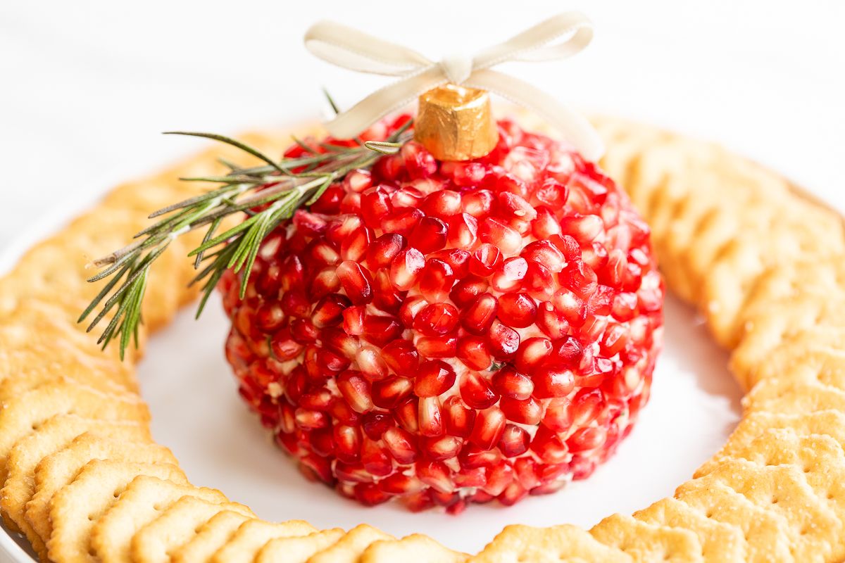 A pomegranate Christmas cheese ball shaped into an ornament, surrounded by crackers on a round white plate. 