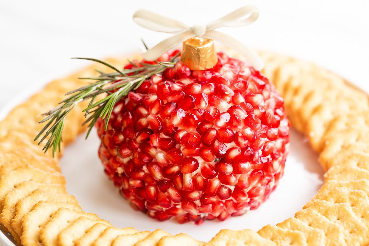 A pomegranate Christmas cheese ball shaped into an ornament, surrounded by crackers on a round white plate.