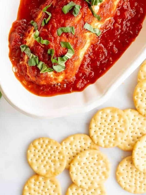 Marinara goat cheese bake on a white platter, crackers and basil surrounding it.
