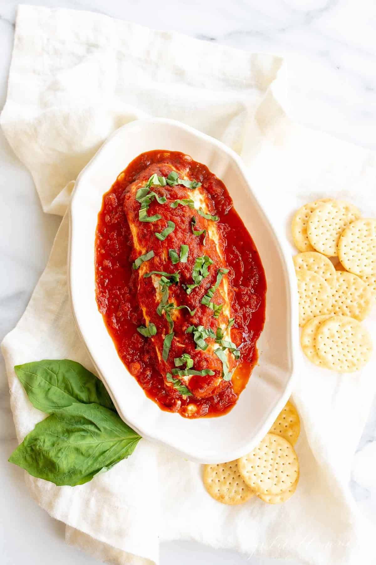 Marinara goat cheese bake on a white platter, crackers and basil surrounding it.