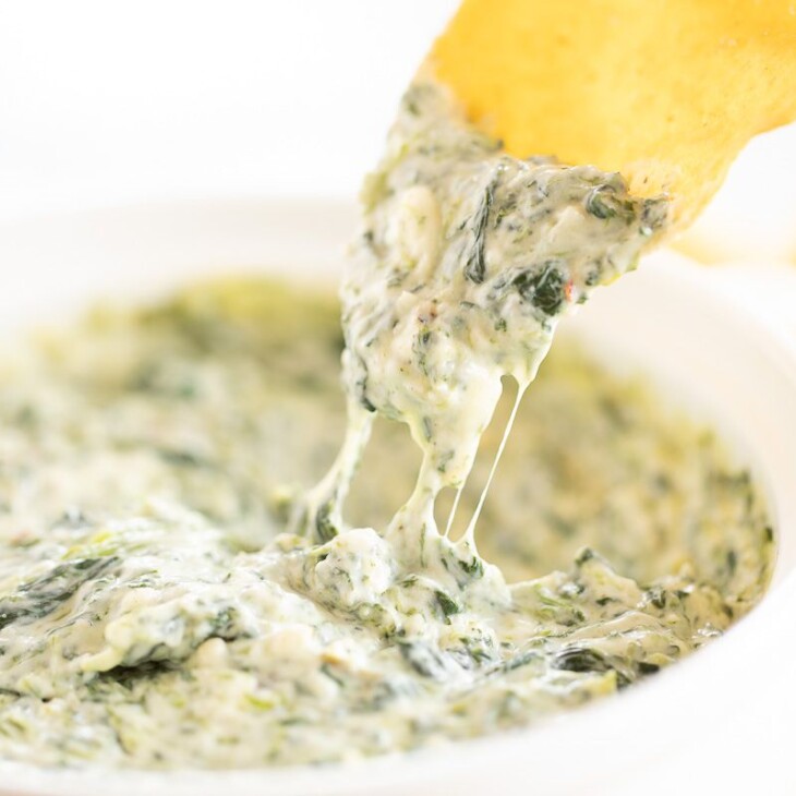 A tortilla chip into a white bowl full of cream cheese spinach dip.