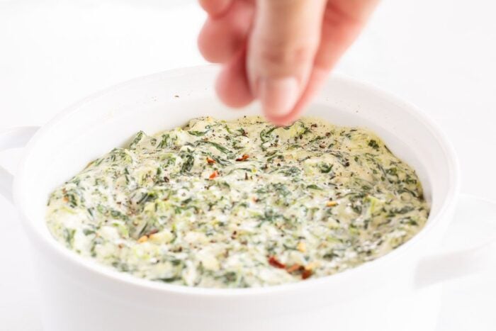 Fingers dipping adding seasoning to a white bowl full of cream cheese spinach dip.