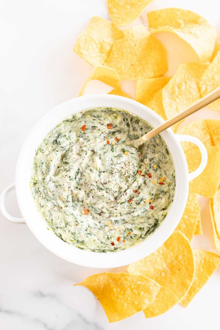 A white bowl full of spinach dip with cream cheese, gold spoon in the bowl and tortilla chips to the side.